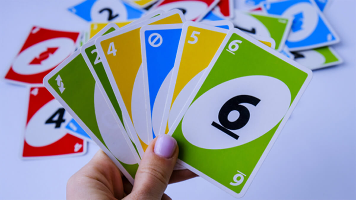 a hand holding a fan of colorful numbered cards against a background scattered with more cards. The cards have different colors with numbers, resembling the gameplay elements of a popular card game, likely used for family or group entertainment.
